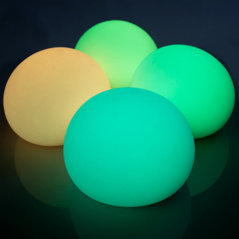 Four Smooshos Jumbo Glow in the Dark Ball glowing balls in yellow, green, and teal on a reflective surface with a dark background