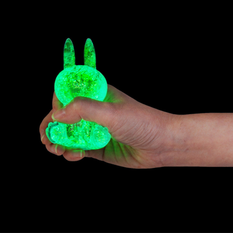 Hand holding a glowing green Smooshos Super Squeeze Glow In The Dark Bunny against a black background