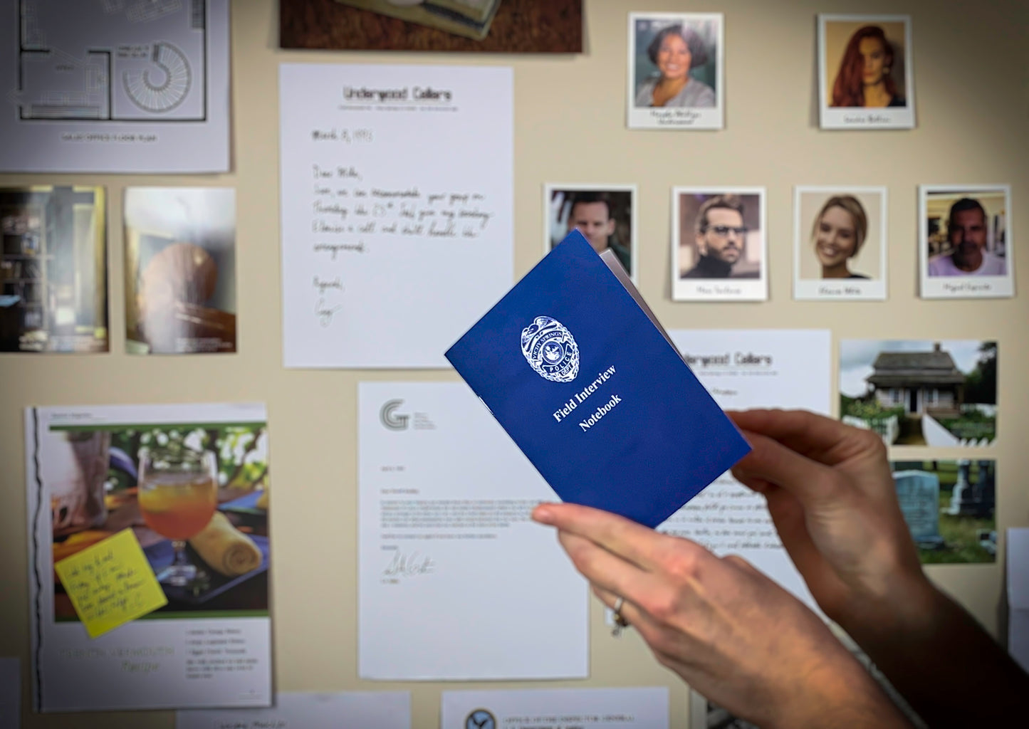 A pair of hands holding a blue "Field Interview Notebook" in front of a beige wall with various documents and photos featured in the murder mystery game Underwood Cellars.