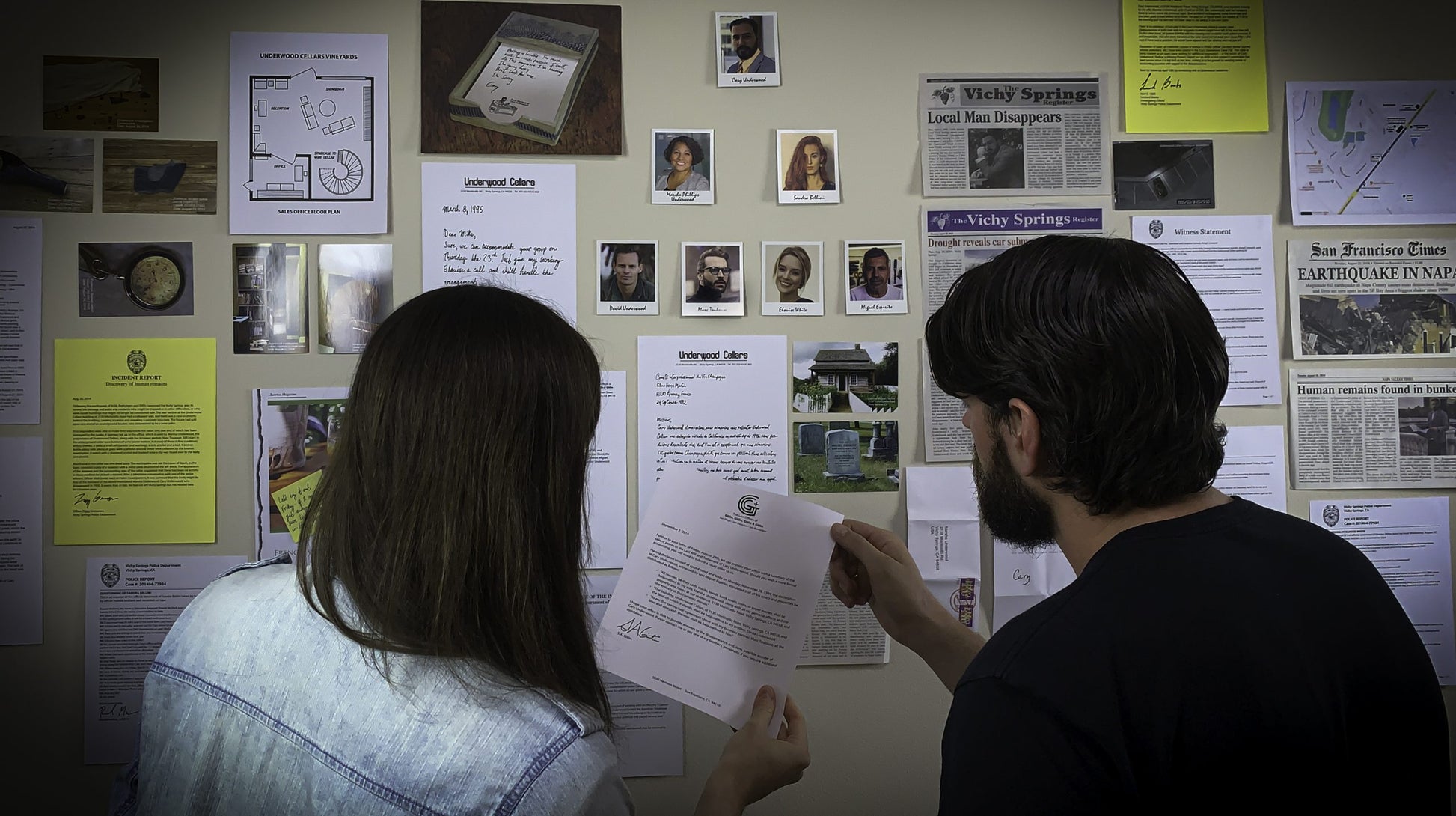 Two people examining documents in front of a wall with various posters and notices from the murder mystery game Underwood Cellars.